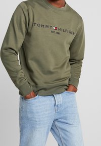 Sudadera verde oliva con cuello redondo, que presenta el texto en azul marino "TOMMY HILFIGER EST. 1985." Combinada con un par de jeans de mezclilla azul claro.
