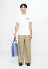 Tricou alb din bumbac cu logo-ul roșu "MARNI", pantaloni largi bej și pantofi negri. Ținând o geantă mare din pânză albastră cu model.