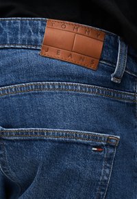 Κοντινή λήψη της πίσω τσέπης από μπλε τζιν με δερμάτινη ετικέτα Tommy Jeans και μικρό κόκκινο, λευκό και μπλε λογότυπο.