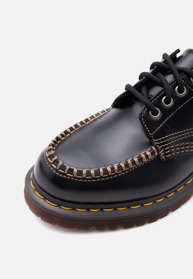 Schwarzer Lederschuh mit dickem braun genähtem Mokassin-Zehenbereich, schwarzen Schnürsenkeln, silbernen Ösen und gelber Naht um die braune Gummisohle.