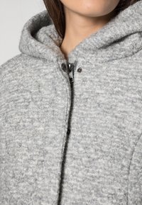 Cappotto in misto lana grigio con superficie testurizzata, ampio cappuccio e chiusura con bottoni. Presenta un design casual realizzato in tessuto morbido.