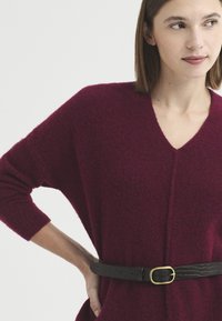 Femme portant un pull violet foncé à col en V avec une ceinture noire à boucle dorée, main posée sur la hanche, regardant sur le côté.