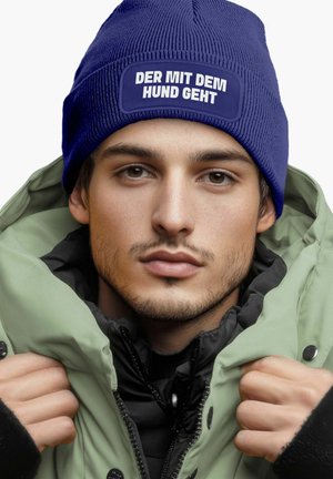 Blue ribbed beanie with a patch featuring white text, worn under a light green puffy jacket with a black inner lining.