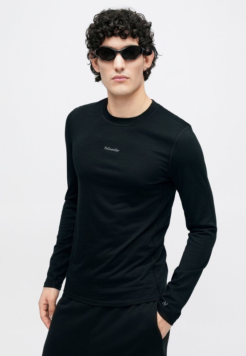 Schwarzes Langarmshirt aus leichtem Stoff, mit rundem Ausschnitt und kleinem Logo auf der Brust. Kombiniert mit sportlichen schwarzen Sonnenbrillen.