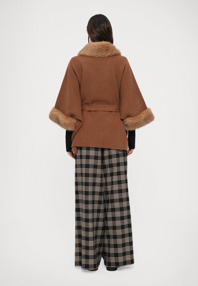 Mujer vestida con una capa recortada de piel marrón y pantalones de cuadros en negro y beige de pierna ancha, de pie de espaldas contra un fondo blanco.