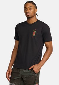 Zwarte t-shirt met korte mouwen van zacht materiaal, met een groene slanggrafiek en een rood accent op de linkerborst. Eenvoudige ronde halslijn.