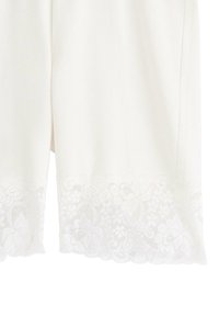 Hvide shorts lavet af blødt stof med en bølget blonderkant med blomster mønstre ved kanten. Glat tekstur og afslappet design.