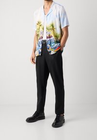 Camisa de manga corta con un gráfico de palmera y hotel en azul, amarillo y verde, combinada con pantalones negros y zapatos negros.