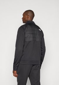 Schwarze Sportjacke mit einem gesteppten Paneel auf dem Rücken, hohem Kragen und gerippten Manschetten. Logo auf dem oberen Rücken angebracht.