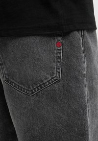 Jeans de mezclilla gris con un bolsillo distintivo que presenta un detalle de costura roja. La tela tiene una textura suave y un corte clásico.