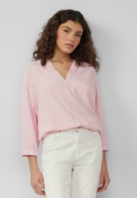 Blouse rose clair à manches longues avec un design en V, fabriquée dans un tissu doux et offrant une coupe ample, associée à un pantalon blanc.