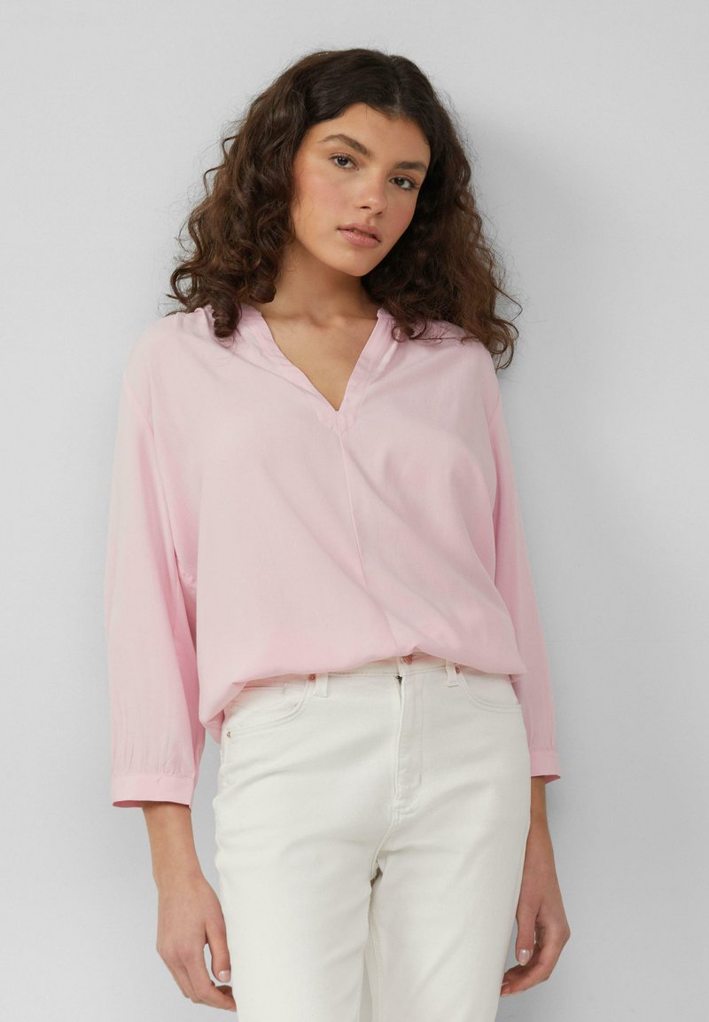 Lichtroze blouse met lange mouwen en een V-hals ontwerp, gemaakt van zachte stof en met een losse pasvorm, gecombineerd met een witte broek.