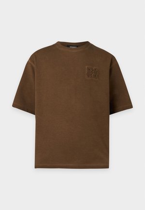Giovane uomo in piedi che indossa una camicia marrone oversize, jeans skinny strappati grigio chiaro, calzini bianchi e scarpe da ginnastica nere con strisce bianche.