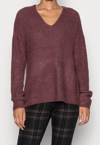 Maglione a V in tessuto ribbed bordeaux con maniche lunghe, vestibilità rilassata e bordi sfrangiati. Abbinato a pantaloni neri a quadri.