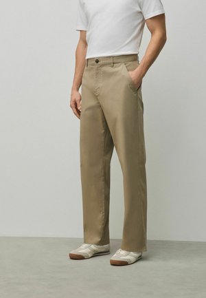 Mann trägt beige gerade geschnittene Hose, weißes T-Shirt und weiße Turnschuhe, steht mit einer Hand in der Tasche.