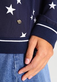 Mörkblå cardigans med vita stjärnmönster, ribbade mudd och en knapp med guldlogo, kombinerad med ett ljusblått denimplagg.