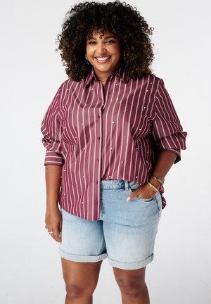 Femme aux cheveux bouclés portant une chemise boutonnée rayée bordeaux avec des perles et un short en jean bleu clair, souriante avec une main dans la poche.