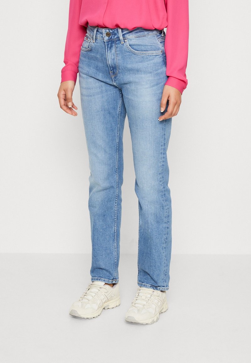 Vaqueros de pierna recta en azul claro con ligero desgastado. Cuenta con cinco bolsillos y costuras sutiles. Combinado con una blusa rosa y zapatillas deportivas ligeras.