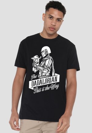 Man wearing black t-shirt with white graphic of Mandalorian holding Baby Yoda and text "The Dadalorian This is the Way."