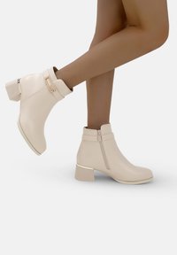 Mujer que lleva botas beige hasta el tobillo con tacones cuadrados y cremalleras laterales, con un pie levantado contra un fondo blanco neutro.