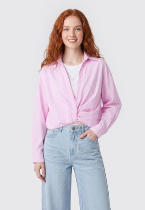 Jonge vrouw met lang rood haar, die een lichtroze blouse met knopen draagt over een wit topje en lichtblauwe spijkerbroek, glimlachend met één hand in de zak.
