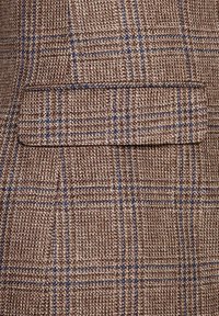 Tissu à carreaux marron et bleu avec une poche à rabat horizontale cousue, montrant un matériau tissé texturé et des détails de couture.
