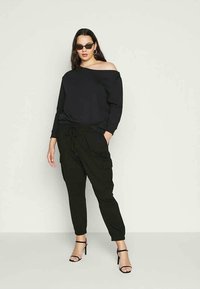 Žena v černé mikině bez ramene, černých cargo kalhotách, černých vysokých sandálech, slunečních brýlích a náušnicích ve tvaru kruhu na neutrálním pozadí.