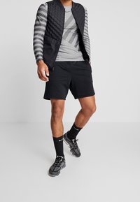 Czarna pikowana kamizelka na szarym longsleeve z poziomymi paskami, czarne szorty i ciemne buty sportowe z warstwową podeszwą.