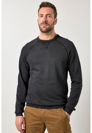 Man staat met een neutrale uitdrukking, draagt een zwart sweatshirt met lange mouwen en bruine cargobroek, tegen een effen achtergrond.