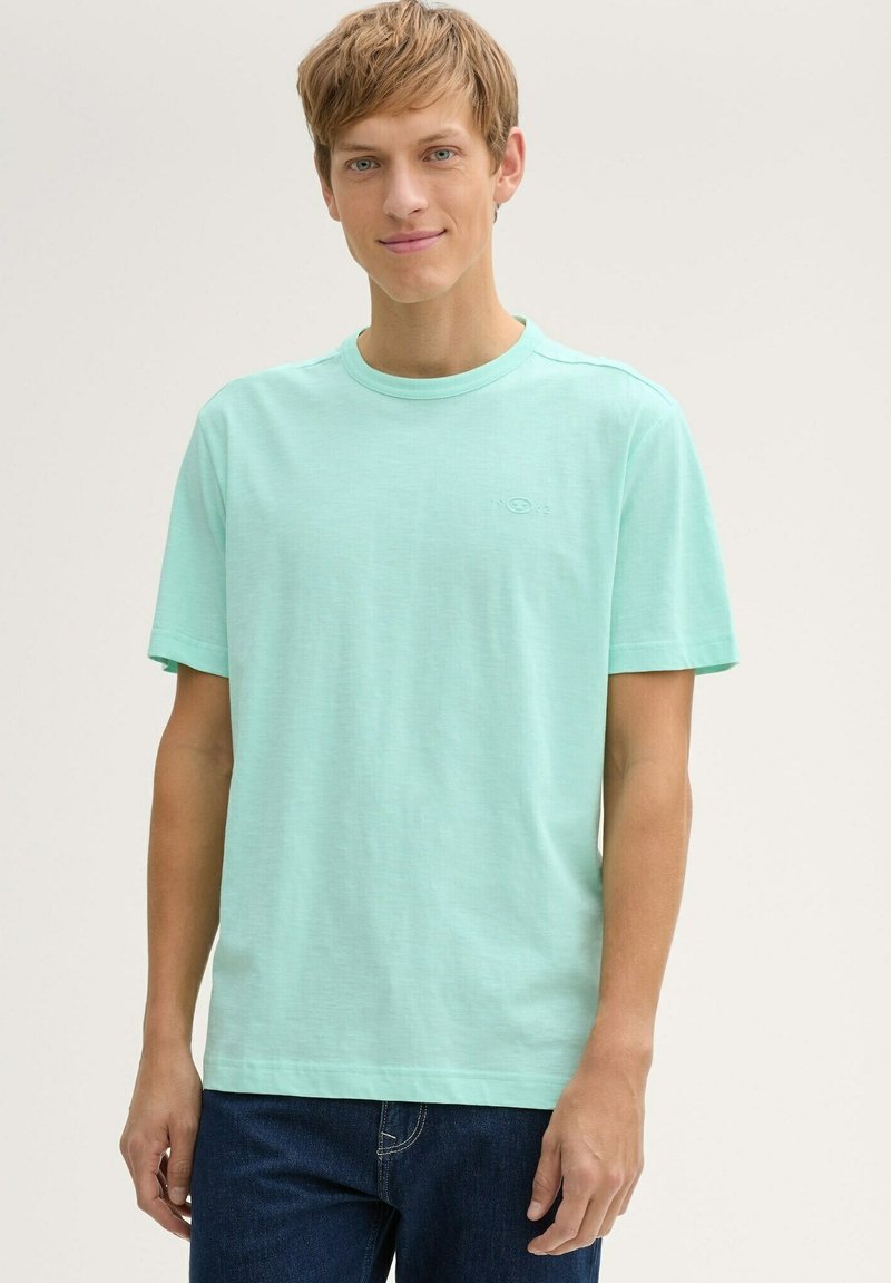 Joven con cabello castaño claro que lleva una camiseta de manga corta verde menta y jeans oscuros, de pie contra un fondo neutro.