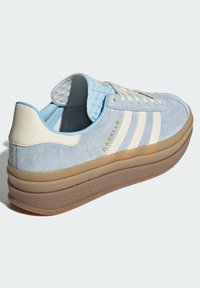 Svetlomodré semišové tenisky s krémovými akcentmi, obsahujúce tri biele pruhy, textúrovaný golier a gumovú podrážku. Zlatofarbené písmo "Gazelle".