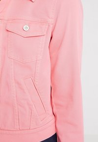 Chaqueta de mezclilla rosa con bolsillos frontales, costuras visibles y herrajes de botón plateados. La textura de la tela es suave con un ajuste ligeramente estructurado.