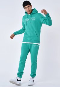 Conjunto de sudadera con capucha y pantalones de chándal en color turquesa, hecho de tela suave, con un bolsillo frontal y detalles en logo blanco. El modelo lleva deportivas blancas.