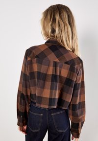 Brown and black plaid button-up shirt with long sleeves and a cropped cut, featuring a back pleat and single-button cuffs.