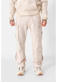 Beige Cargo-Hosen aus Baumwolle mit mehreren Taschen, einer lockeren Passform und gefütterten Säumen, kombiniert mit hellen Sneakern.