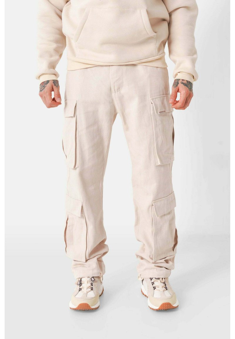 Beige Cargo-Hosen aus Baumwolle mit mehreren Taschen, einer lockeren Passform und gefütterten Säumen, kombiniert mit hellen Sneakern.