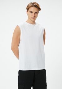 Vit ärmlös t-shirt i mjukt bomullstyg med rund hals och avslappnad passform, ihopparad med svarta cargobyxor.