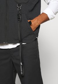 Gilet noir texturé avec une fermeture éclair à l'avant, doté d'un détail de clip et de sangle. Porté sur une chemise blanche, associé à un pantalon noir arborant un logo.