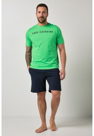 Uomo con braccio tatuato che indossa una maglietta verde brillante con la scritta "LAKE CAVEDINE" e pantaloncini blu scuro, in piedi a piedi nudi su uno sfondo chiaro.
