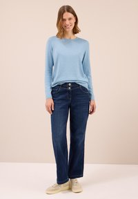 Hellblaues Langarmpullover kombiniert mit dunkelblauen weit geschnittenen Jeans. Die Jeans besitzen Fronttaschen und einen Zwei-Knopf-Verschluss. Cremefarbene Sneakers.