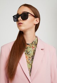 Ochelari de soare negri cu o ramă lucioasă și accente roșii. Purtați cu un sacou roz pastel și o bluză verde cu un model. Păr lung și castaniu.
