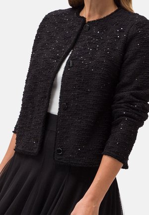 Cardigan texturé noir avec petites paillettes et boutons noirs porté sur un haut blanc, associé à une jupe plissée noire.