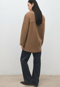 Cappotto di lana marrone con silhouette squadrata, maniche ampie e orlo dritto, abbinato a jeans scuri svasati e scarpe marroni.