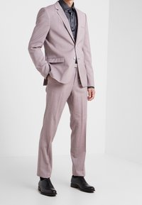 Abito di un leggero viola con un motivo testurizzato, giacca monopetto, pantaloni coordinati e scarpe eleganti nere, abbinato a una camicia scura.