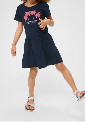 Niña con vestido azul marino de capas, con un gráfico de palmeras rosas y el texto "La felicidad viene con vibras tropicales", posando con una pierna extendida.