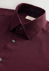 Cămașă texturată bordeaux cu guler, plachetă butonată, butoane gri și o etichetă branduită în interiorul gulerului.