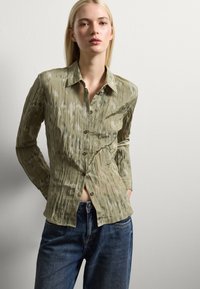 Blusa verde chiaro, con texture, bottoni frontali, maniche lunghe e un motivo sottile; abbinata a jeans in denim blu.