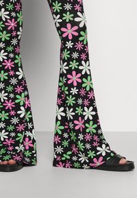 Uitlopende zwarte broeken met een roze, groene en witte bloemenpatroon, gedragen met zwarte sandalen met open neuzen die de tenen tonen.