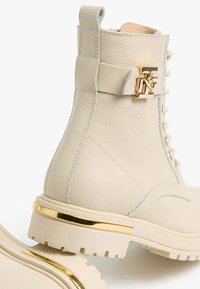 Bottines en cuir beige avec ferrures dorées, bout rond, semelle épaisse et détail de couture contrastée, avec une bride ornée d'un logo.