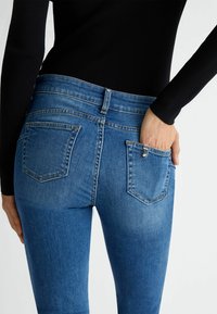 Persona vestida con jeans azules que tienen bolsillos traseros, una mano descansando dentro del bolsillo trasero derecho y una blusa negra de manga larga.